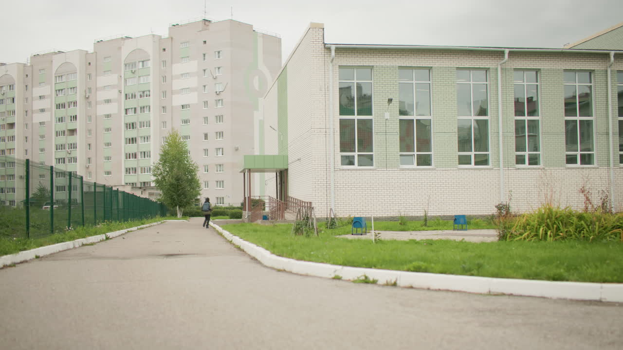 Vista trasera de un niño con mochila corriendo hacia un salón de actos rodeado de vegetación y altos edificios residenciales con estructuras de hierro visibles a lo largo del camino bajo un cielo nublado