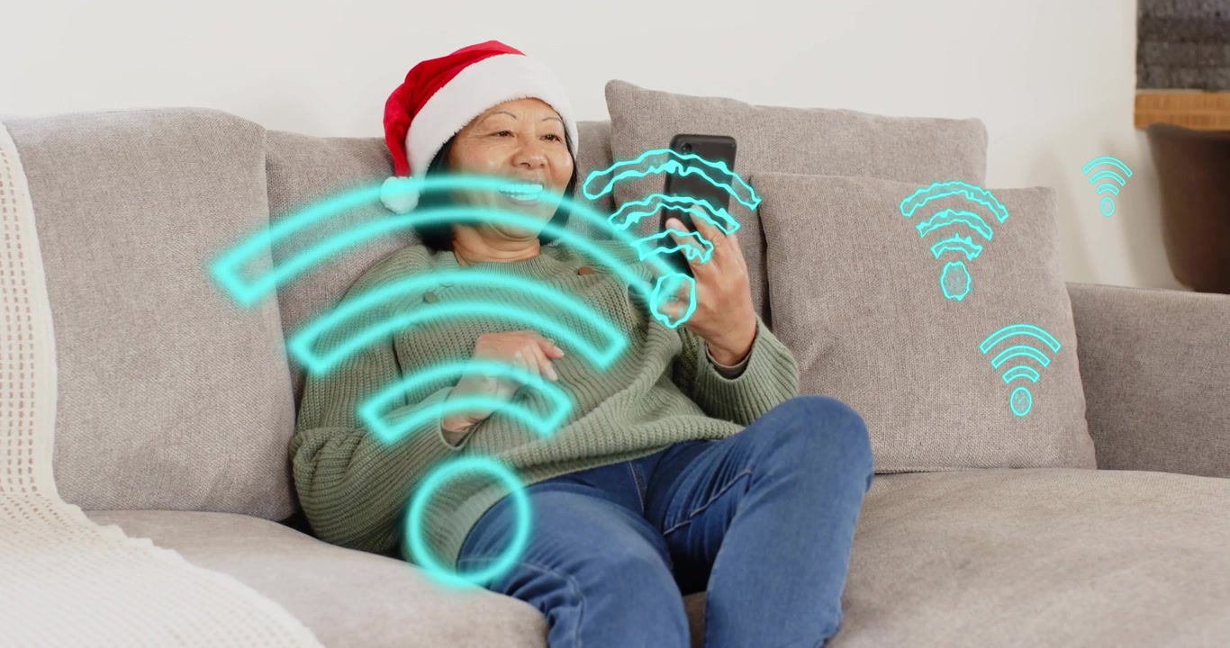 En ældre kvinde med nissehue vifter med sin smartphone på sofaen, hvilket genererer Wi-Fi-ikoner til en julechat.
