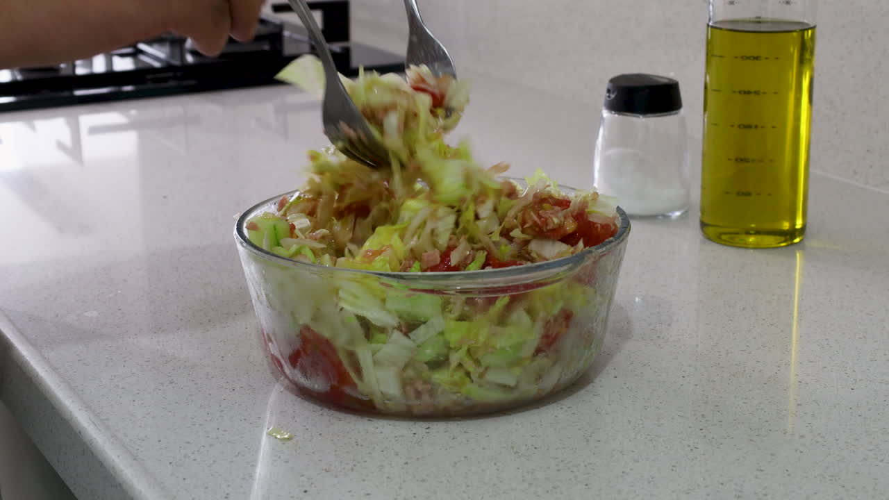Close-up of a person mixing fresh cabbage, tomato, and tuna salad in a glass bowl, highlighting healthy homemade meal preparation in the kitchen