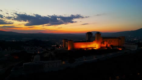 Luftaufnahme-Der-Beleuchteten-Festung-Rocca-Albornoziana-Mit-Sonnenuntergangshimmel-Im-Hintergrund