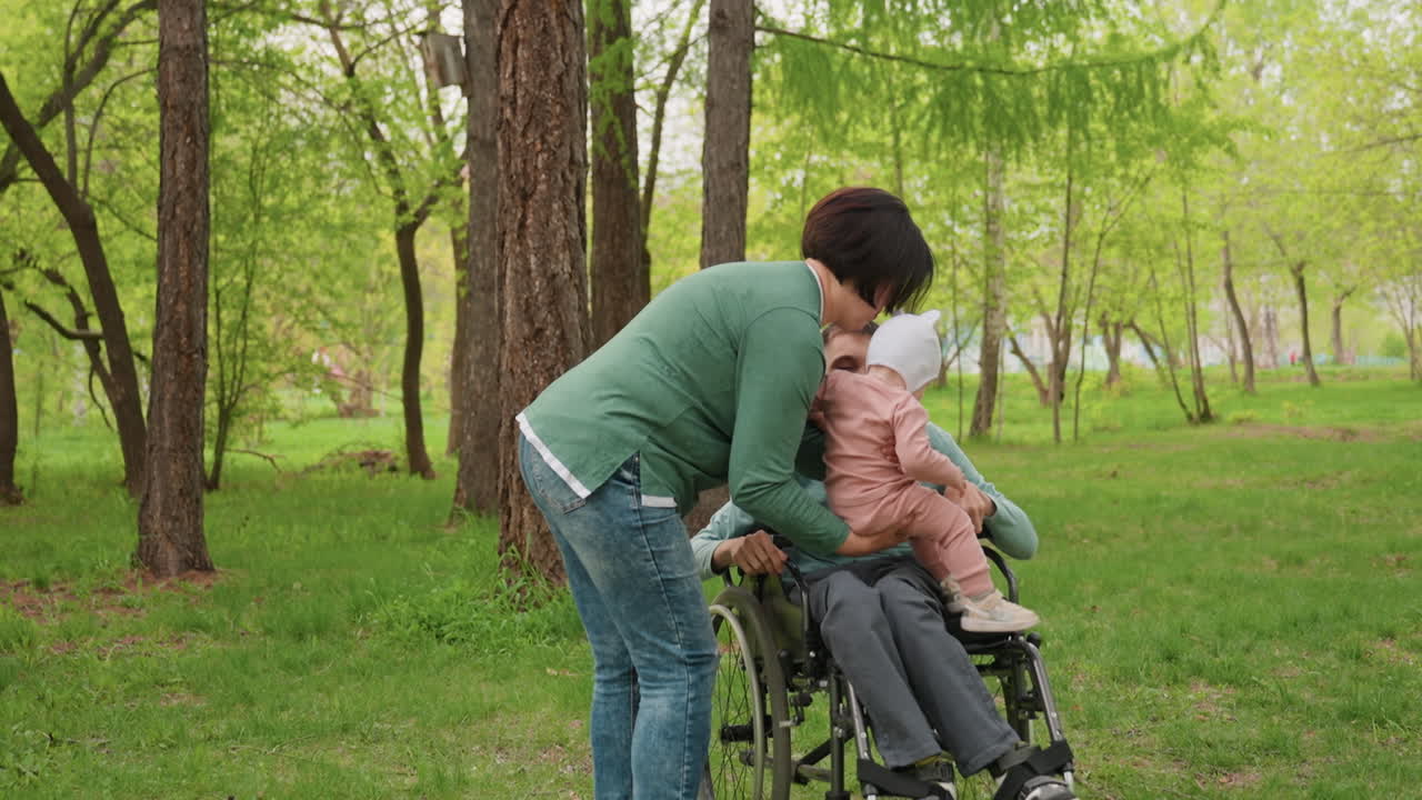 Wide Shot White Father In Wheelchair Meeting Baby Held By Mother, Inclusive Family Scene In Park, Gentle Interaction, Accessible Path, Supportive Spouse Assisting, Warm Community Atmosphere