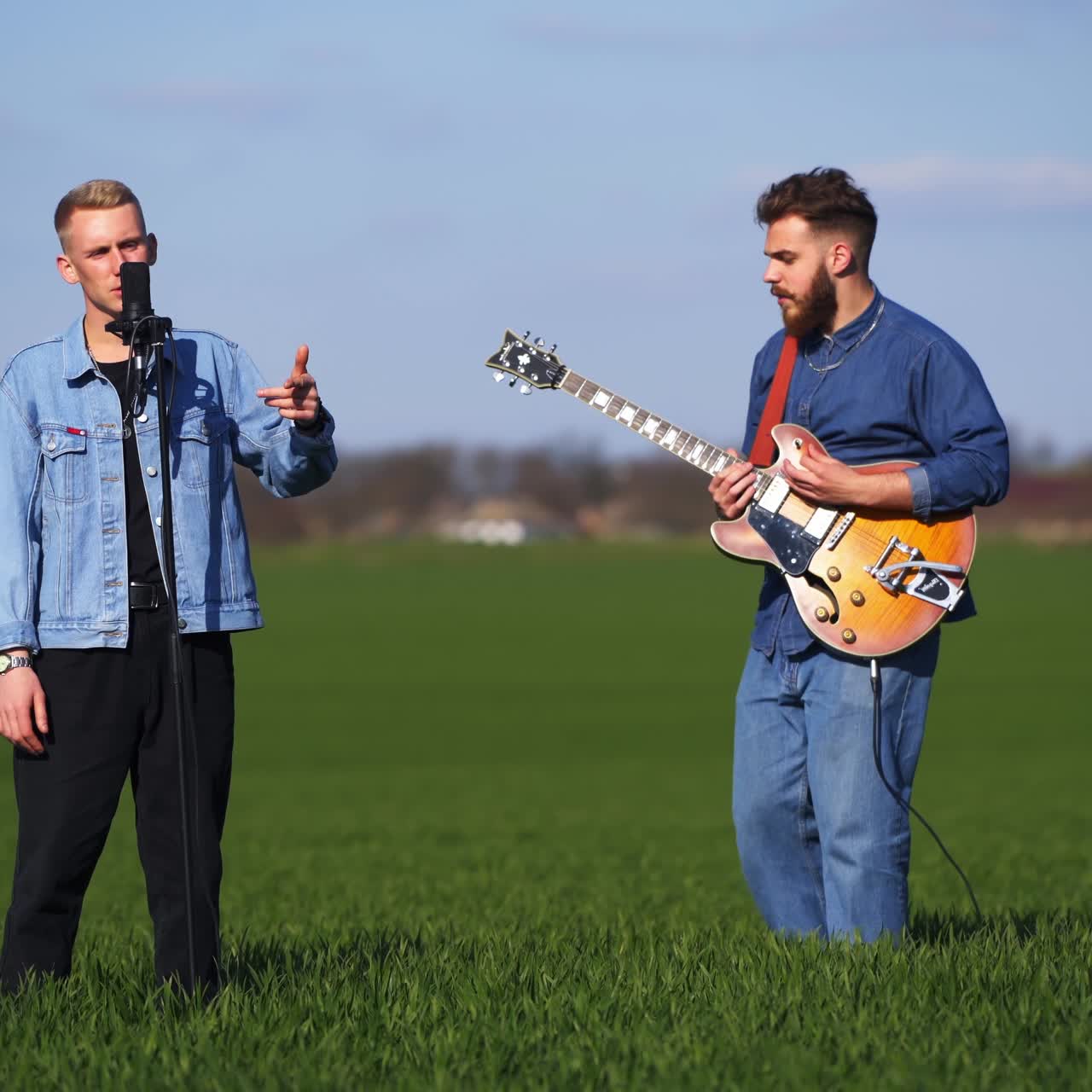 Cantante masculino cantando con música de guitarra interpretada por un músico barbudo. Dos miembros de una banda musical actuando con un campo verde de fondo.