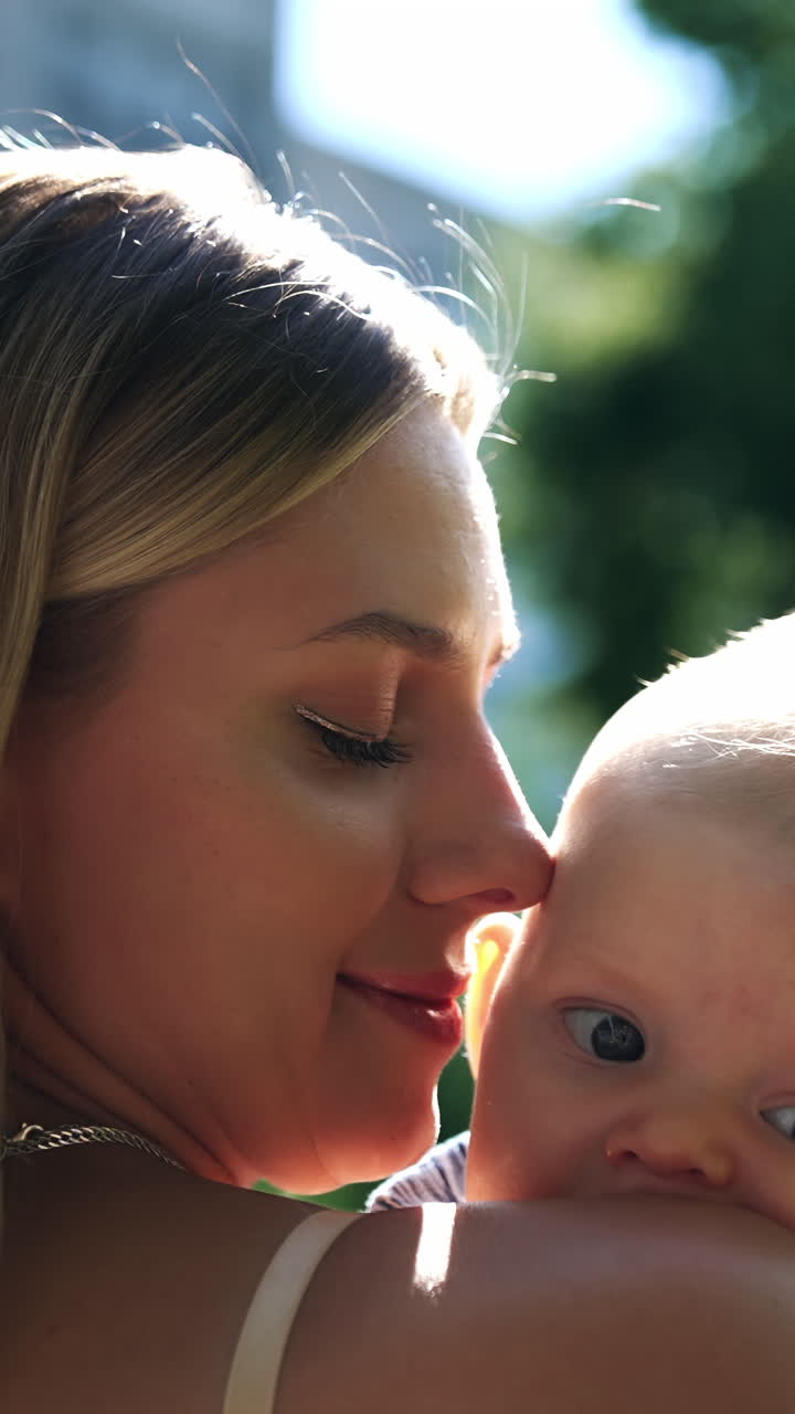 Beautiful blonde woman stands half-back to camera holding a baby. Loving mom caressing her son tenderly. Close up. Vertical video