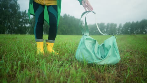 lower view of person in yellow rain boots and leggings dropping a mint-colored bag on grassy field, the background features an expansive grassy field and trees under a cloudy sky