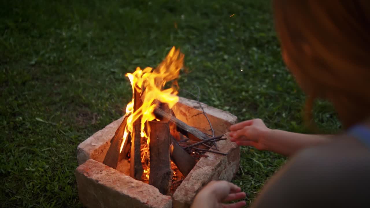 Donna che mette piccoli bastoncini in un falò acceso in un focolare sull'erba, mantenendo la fiamma e catturando l'attività di campeggio all'aperto e l'ambiente naturale a Savci, Slovenia.