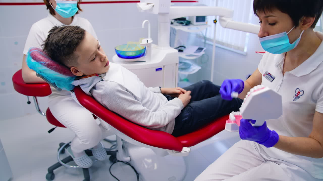 Pediatric dentistry. Boy is sitting in dental chair and listens to the doctor in dental clinic. Female stomatologist showing the child how to brush teeth on a jaw mock.