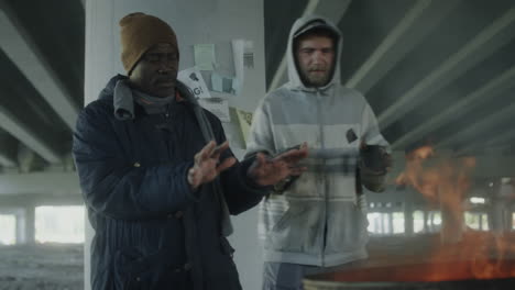 homeless men warming themselves by a fire under a bridge
