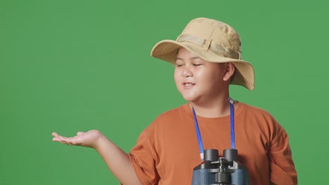 asian tourist boy with a hat and binoculars smiling and pointing to side while standing on the green screen background. boy researcher examines something, travel tourism adventure concept, close up