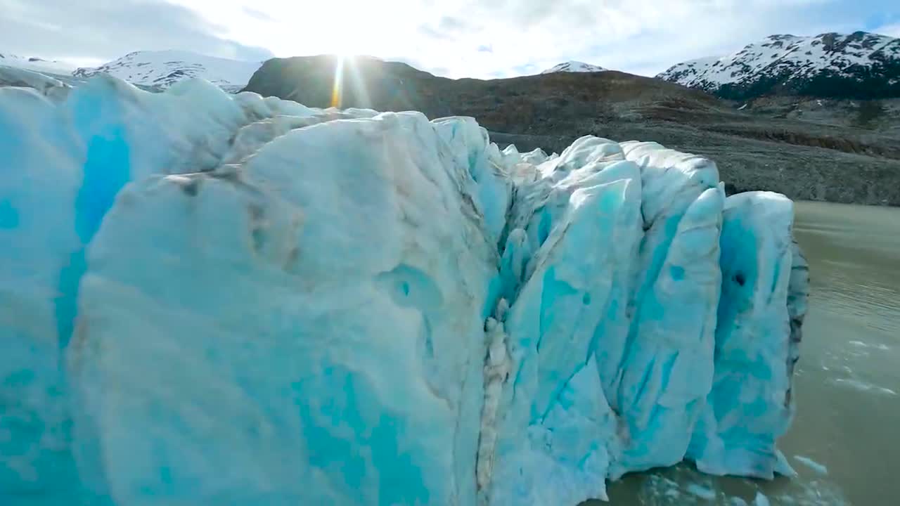 FPV aerial drone flying close to icy blue glaciers and in between them on a sunny day between Greenland or Iceland mountains that are covered with some snow. The Glaciers are in brown ocean water.