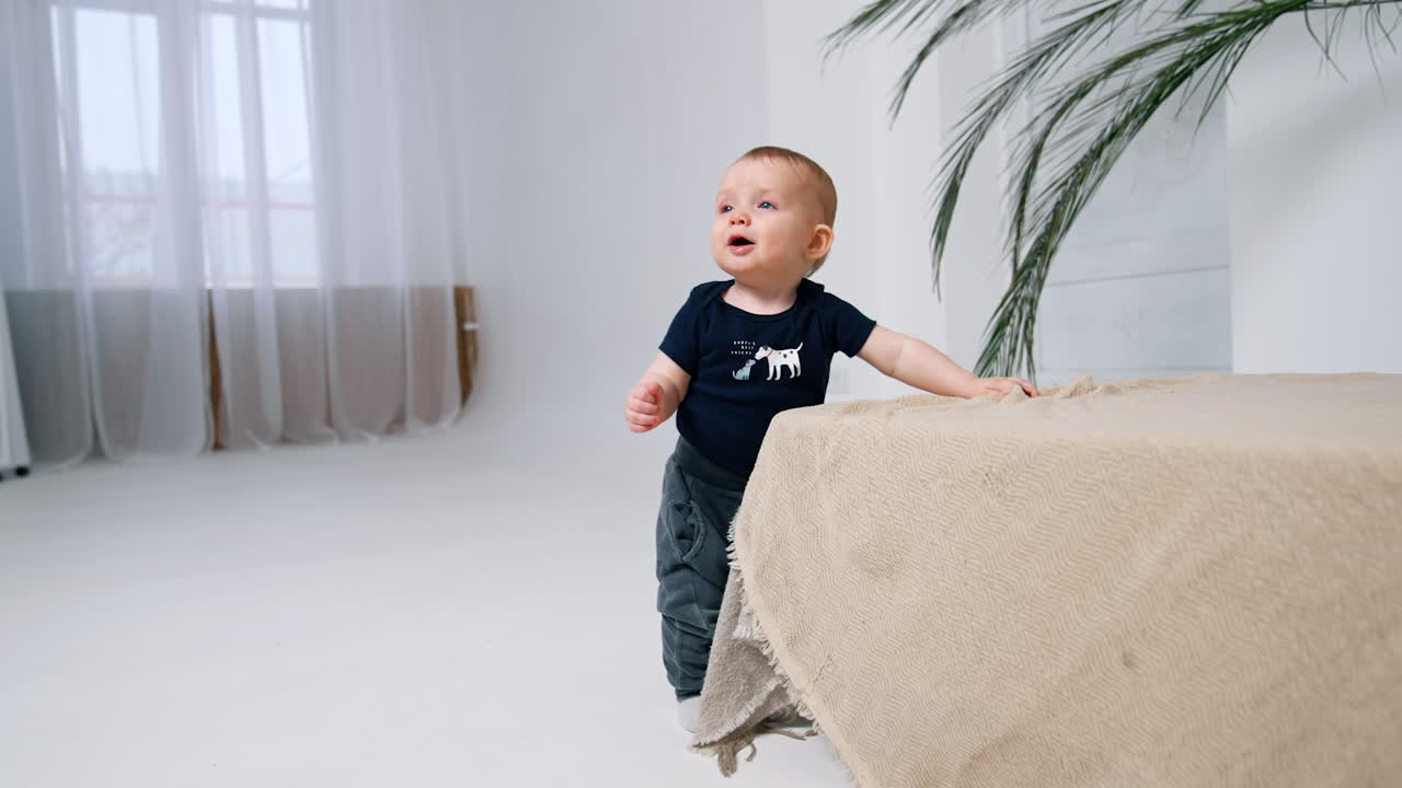 Un niño adorable y sonriente está de pie cerca de la cama, mirando hacia arriba. Un bebé pequeño y lindo baja al suelo.