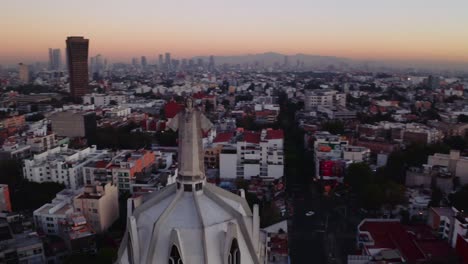 Tomas-Dinámicas-De-Drones-En-Picada-Amplia-Del-Estado-De-Jesús-Masivo-Que-Se-Eleva-Sobre-La-Ciudad-De-México-Al-Atardecer,-Con-La-Parroquia-Del-Purismo-Corazon-De-Maria-Y-Edificios-En-La-Parte-De-Atrás