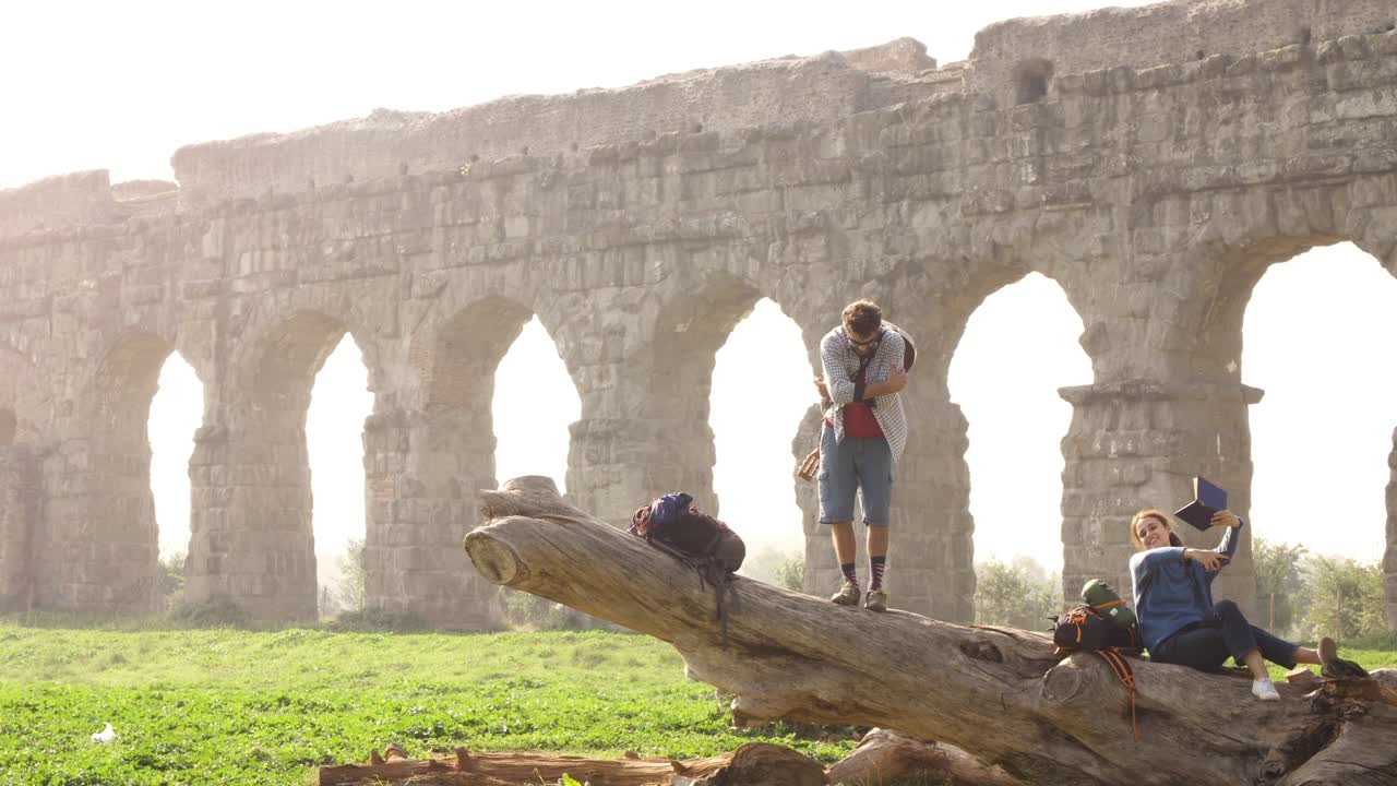 Gelukkig jong stel rugzaktoeristen met gitaar op een boomstam poseert voor een foto voor de ruïnes van een oud Romeins aquaduct in het romantische Parco degli Acquedotti park in Rome bij mistige zonsopgang, slow motion.