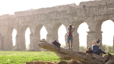 happy young couple backpackers tourists with guitar on a log trunk posing for picture in front of ancient roman aqueduct ruins in romantic parco degli acquedotti park in rome at misty sunrise slow motion