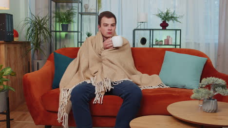 sick teen man wear hat wrapped in plaid sit alone shivering from cold on sofa drinking hot tea