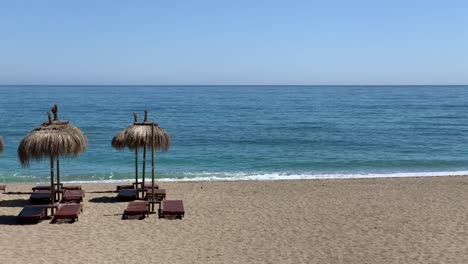 Toma-Estática-De-La-Playa-Con-Sombrillas-Estilo-Tiki-Tropical-En-Una-Soleada-Costa-De-Verano-En-Marbella,-Malaga,-España,-Costa-Del-Sol