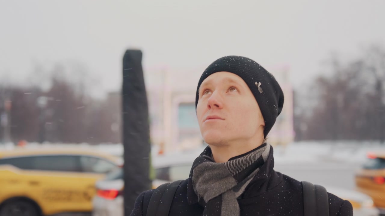 Primo piano di un uomo che indossa abiti invernali, in piedi all'aperto sotto la neve che cade, con taxi gialli e uno sfondo urbano. Il soggetto rimane immobile mentre sono visibili una sciarpa grigio chiaro e un cappotto nero.