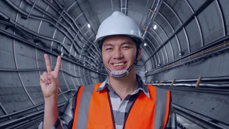 close up of asian male engineer with safety helmet smiling and showing peace gesture while standing in underground subway tunnel