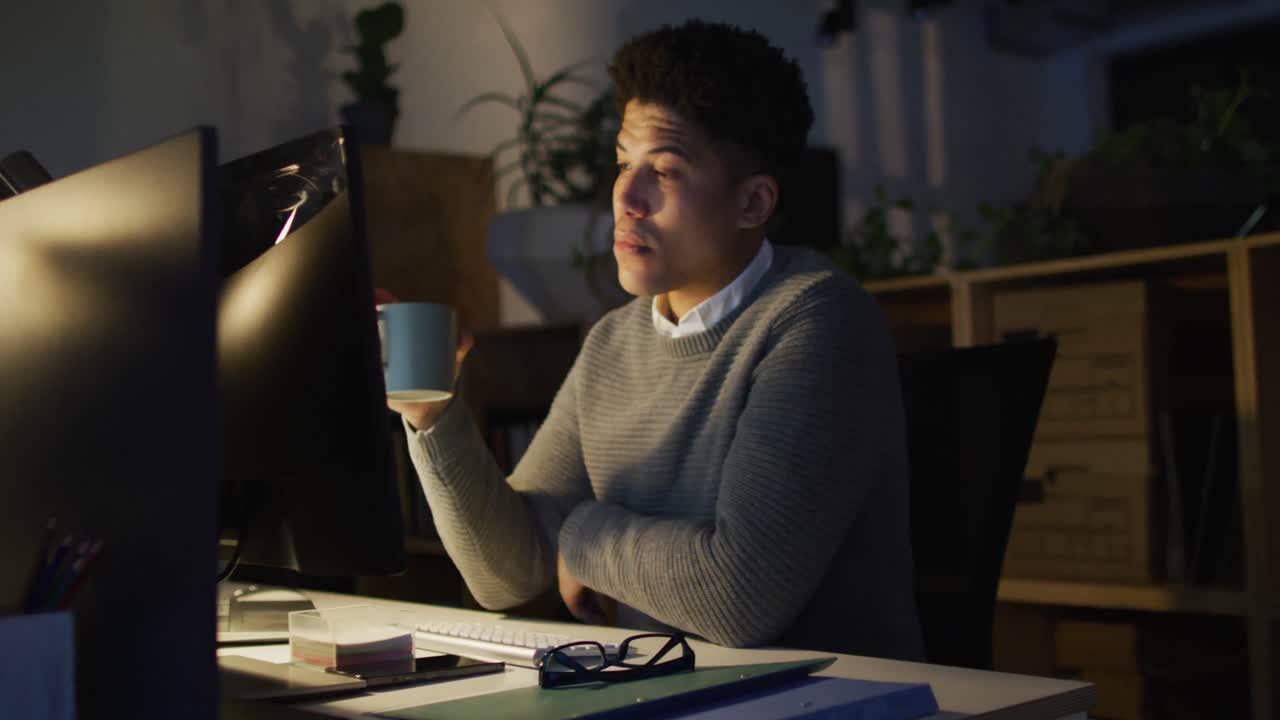 Video of tired biracial man with coffee using computer, working late in office