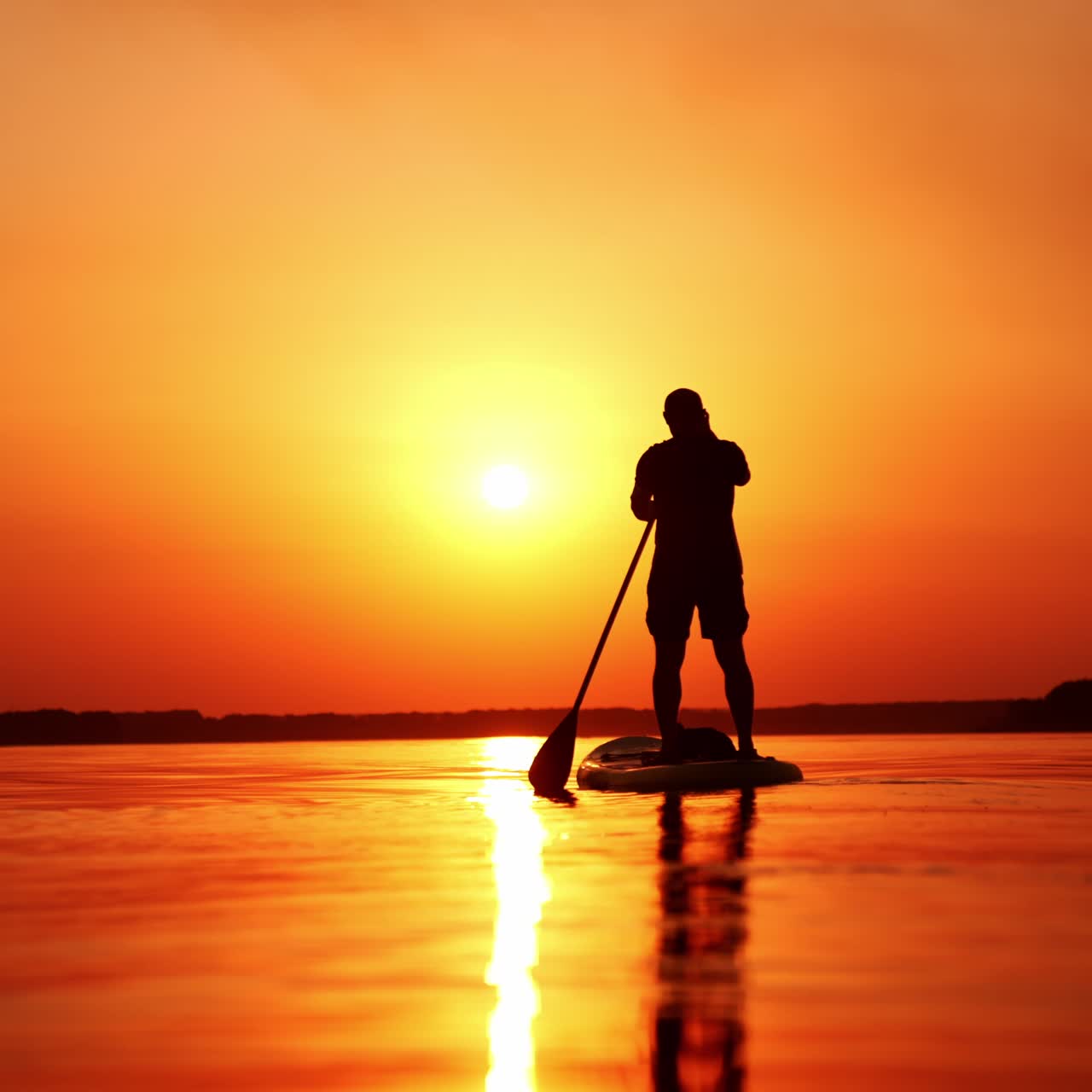 Male silhouette wearing t-shirt and shorts standing on sup board. Man floating by the river rowing with a paddle. Orange sunset backdrop. Low angle view