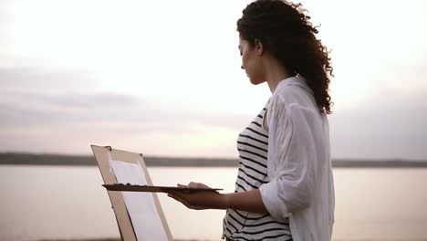 Beautiful curly girl draws a picture on the meadow near the lake using a palette with paints and brush. Easel and paper. Side view