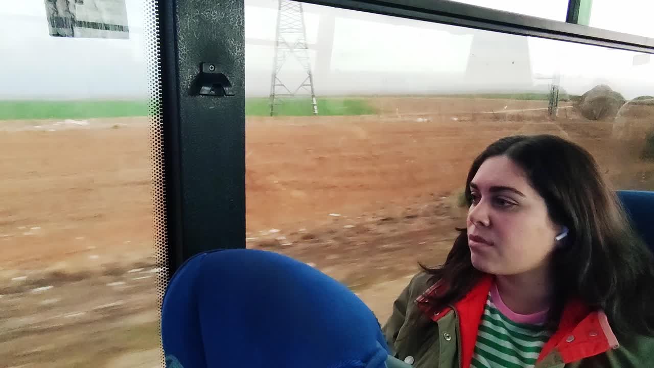 A woman is sitting on a bus with her earbuds in. The bus is on a road with a lot of empty space around it