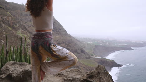 Slow-motion-reveals-woman-meditating-in-yoga-warrior-pose-amid-ocean,-beach,-and-rock-mountains,-representing-motivation,-inspiration,-and-harmonious-blend-of-fitness-and-nature