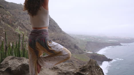 slow motion reveals woman meditating in yoga warrior pose amid ocean, beach, and rock mountains, representing motivation, inspiration, and harmonious blend of fitness and nature