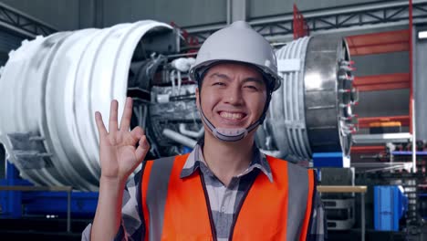 close up of asian male engineer with safety helmet smiling and showing okay gesture to the camera while standing with airplane engine maintenance conducted, analytics checking the turbine