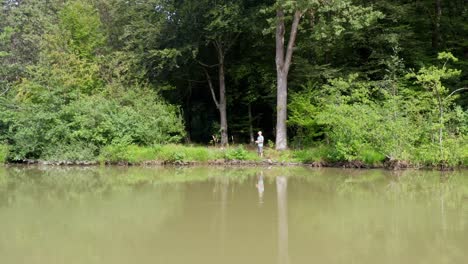 Male-angler-stands-on-shore-of-murky-lake-and-reels-in-his-cast-fishing-line,-approaching-drone-shot