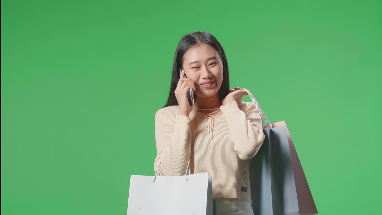 An Asian Shopping Woman With Shopping Bags Answering The Call And Talking On Mobile Phone While Standing In Front Of Green Screen