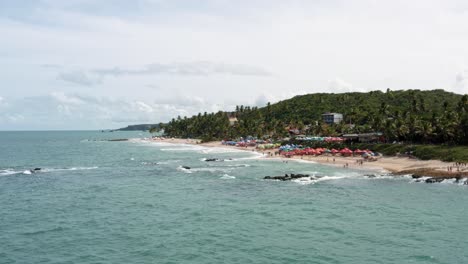 Dolly-in-aerial-drone-shot-of-the-popular-tropical-Coquerinhos-beach-surrounded-by-palm-trees-and-covered-in-umbrellas-and-small-waves-crashing-into-exposed-rocks-in-Conde,-Paraiba,-Brazil-in-summer