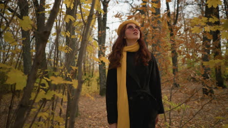 stylish lady in yellow beret and muffler walks through a forest path, looking around as autumn leaves sway gently with the wind, immersed in the tranquil beauty of the season