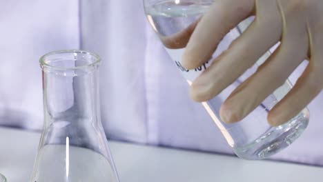 a gloved hand pours liquid from a bottle into a labeled flask beside a test tube.