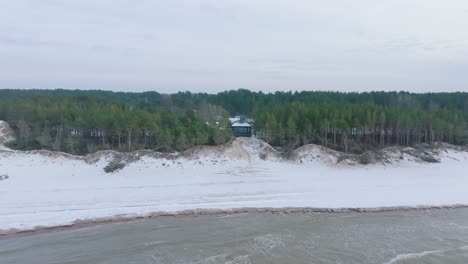 aerial establishing view of baltic sea coast on a overcast winter day, a tiny gray holiday house at the beach with white sand, coastal erosion, climate changes, wide drone shot moving forward