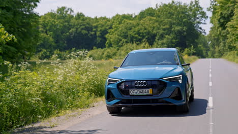 beautiful e-tron audi moving on empty road. sportive electric car of light blue color driving slowly on green nature background. front view.