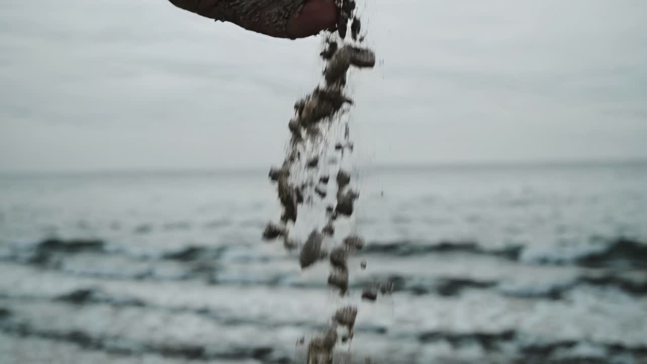 Close-up video shot of a hand releasing sand, captured from a low angle, with a blurred ocean