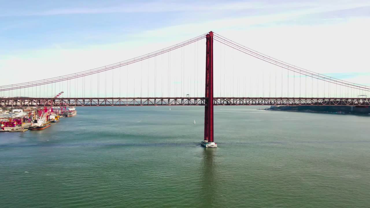 Aerial drone time lapse or hyperlapse video of Ponte 25 de Abril red colored bridge going over Tagus river or Atlantic ocean at sunny day. Cars, vehicles and trains crossing on the bridge fast. Large