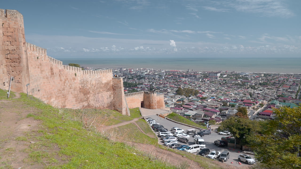 Mura di una fortezza che si affacciano sullo skyline di una città costiera, collina erbosa con auto parcheggiate e strada tortuosa, mare aperto. Creativo