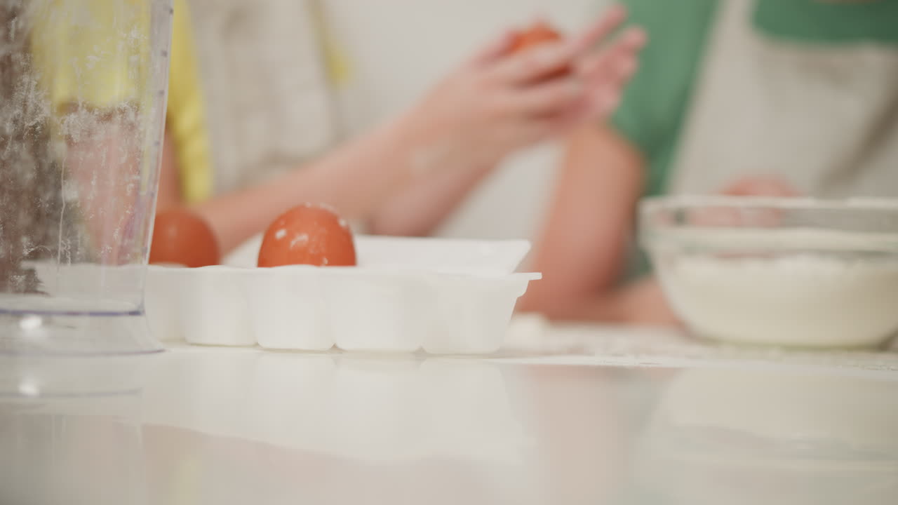 close up of flour-covered child's hand reaching into white egg carton to pick egg during baking session, kitchen tools and glass bowl with flour in background
