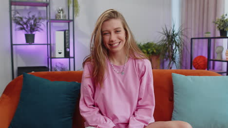 young woman smiling friendly at camera, waving hands gesturing hello, hi, greeting at home on couch