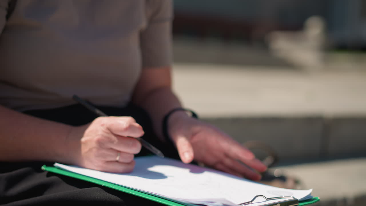 Close up of woman writing on clipboard outdoors, gentle wind lifting page, sunlight highlighting hands and pen, capturing focus, calm atmosphere, and quiet moment of concentration