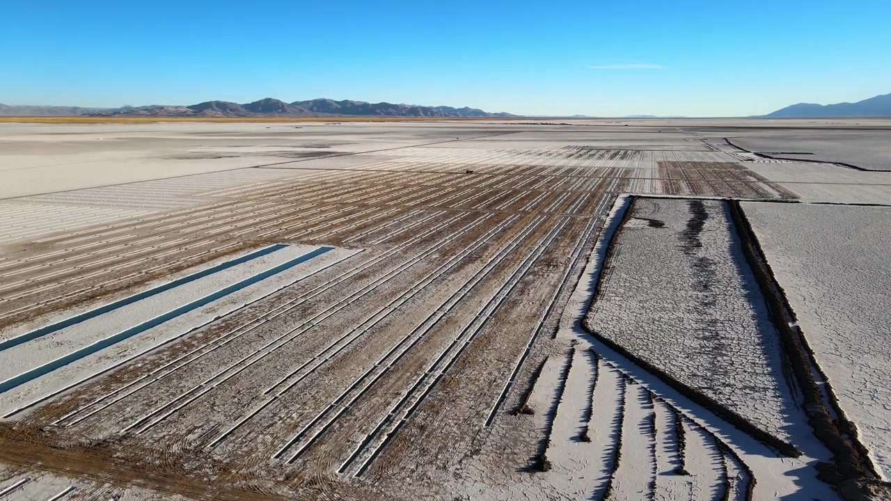 Salt extraction grids and shallow evaporation ponds cut across the white Salinas Grandes salt flats in Jujuy Province under clear blue sky, with distant Andes mountains on the horizon.
