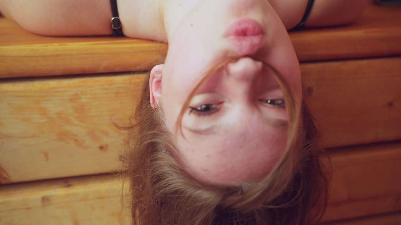 White kid resting her head backward on wooden bench, holding hair with her nose, eyes closed, enjoying relaxing moment, sauna environment creates a peaceful and soothing atmosphere