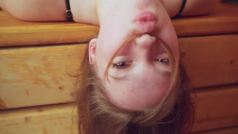 white kid resting her head backward on wooden bench, holding hair with her nose, eyes closed, enjoying relaxing moment, sauna environment creates a peaceful and soothing atmosphere