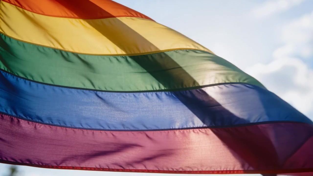 Waving rainbow pride flag letting sun glare through thin fabric under open sky, wind causing waves
