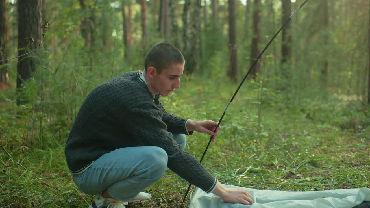 Boy squats in forest setting as he holds flexible tent pole and secures fabric to ground, preparing for outdoor camping with rising movement and surrounding nature bathed in soft daylight glow
