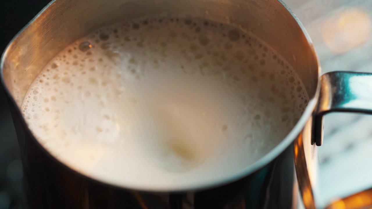 Vista em close-up de uma jarra de metal cheia de leite vaporizado, mostrando a textura cremosa e as bolhas na superfície, perfeita para o preparo de café e bebidas.