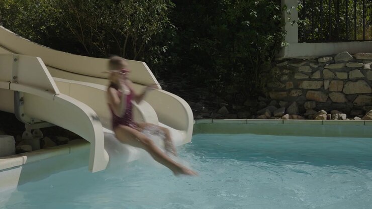 Girl slides down a white water slide into a swimming pool, splashing water, slow motion, wearing swimming goggles, outdoor pool surrounded by green bushes and a stone wall