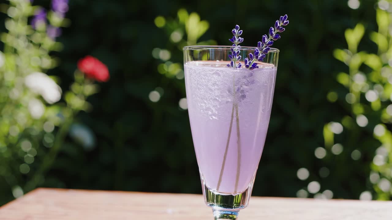 Glass filled with a refreshing homemade lavender cocktail, featuring ice and delicate lavender flowers, resting on a wooden table amidst a blooming summer garden