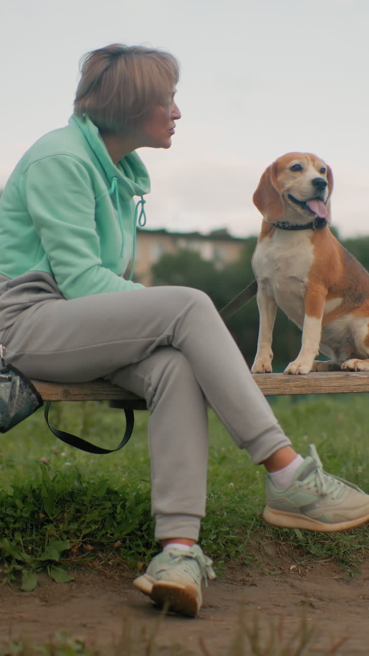 잔디가 있는 공원에서 저녁 무렵 민트색 후드티와 회색 조깅 바지를 입은 백인 여성이 벤치에 앉아 목줄을 한 비글을 부드럽게 쓰다듬으며 다정한 눈빛으로 교감하는 편안한 야외 분위기